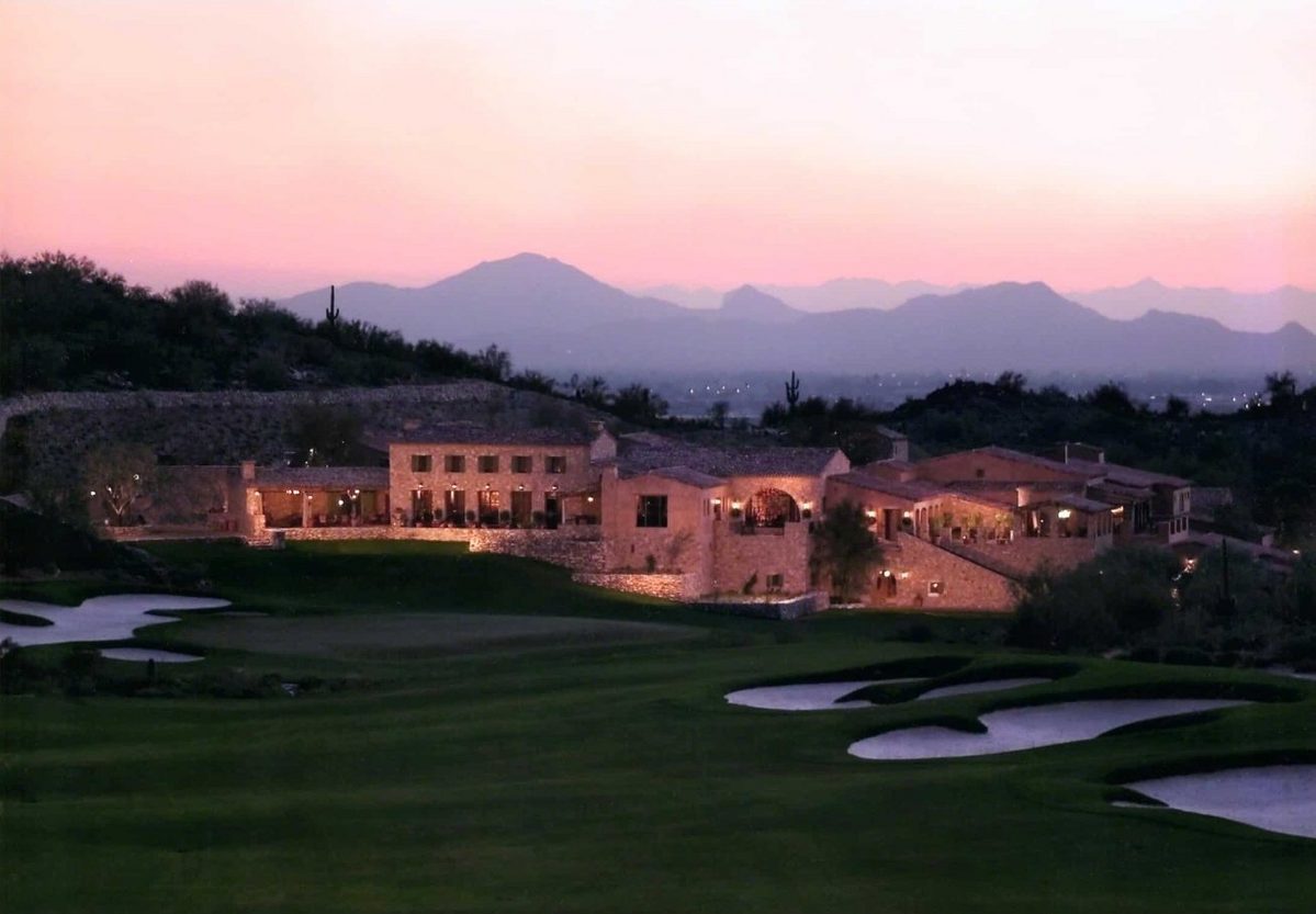 The Silverleaf Club clubhouse at twilight with its windows lit, seen across the dark fairways and pale bunkers under a pink evening sky